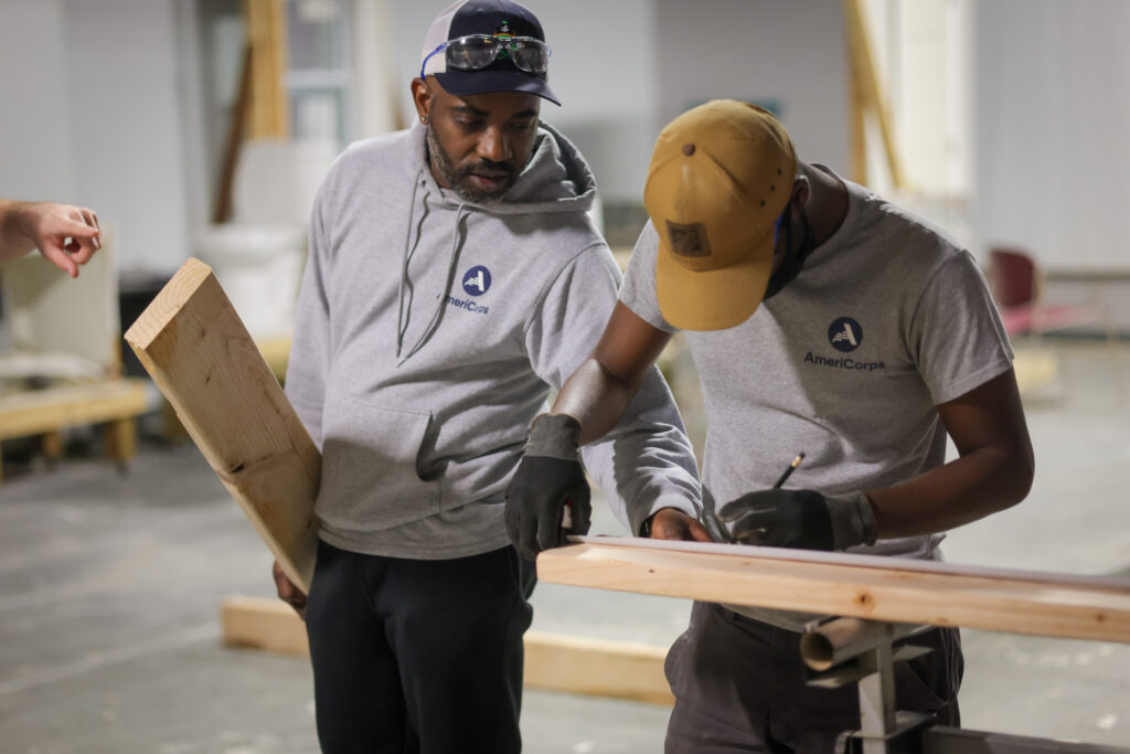 A middle-aged Black man wearing protective glasses helps a younger Black man mark a piece of wood for cutting